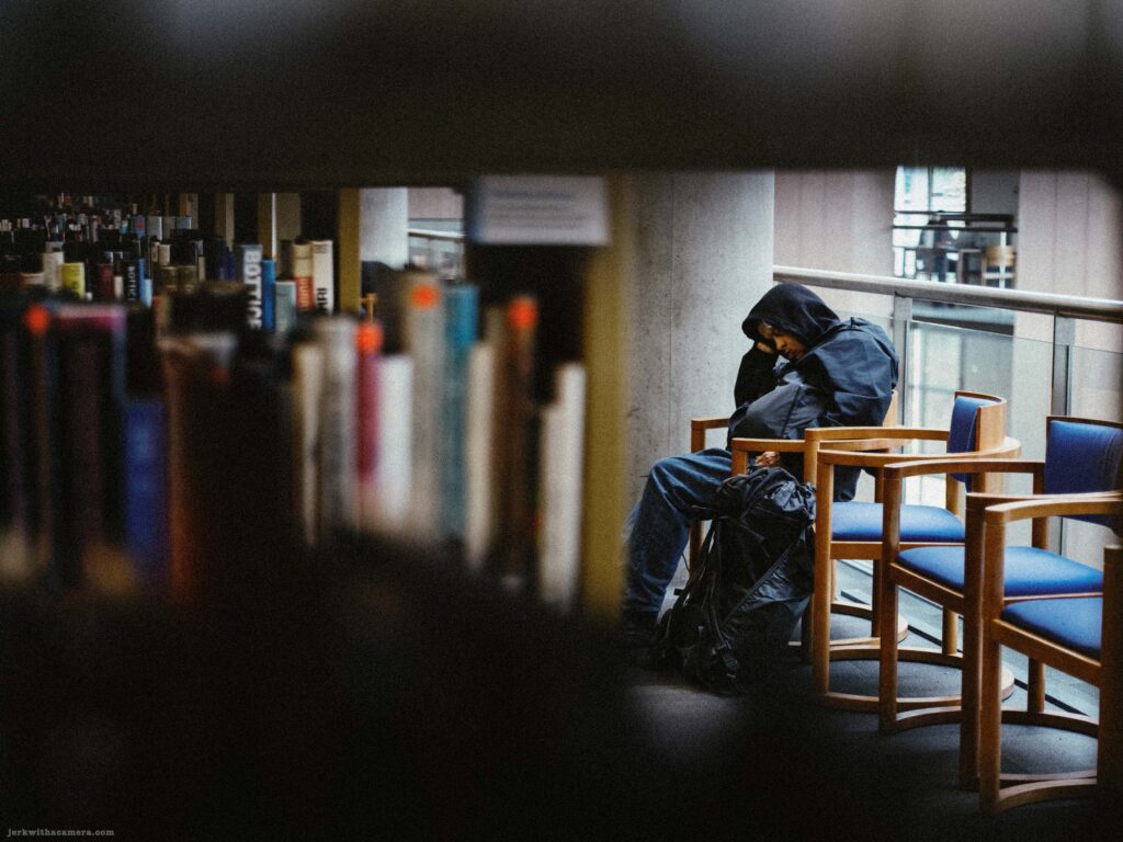 Wider view of person slumped in library chair between bookshelves. More books visible on shelves. Second person faintly visible in background.