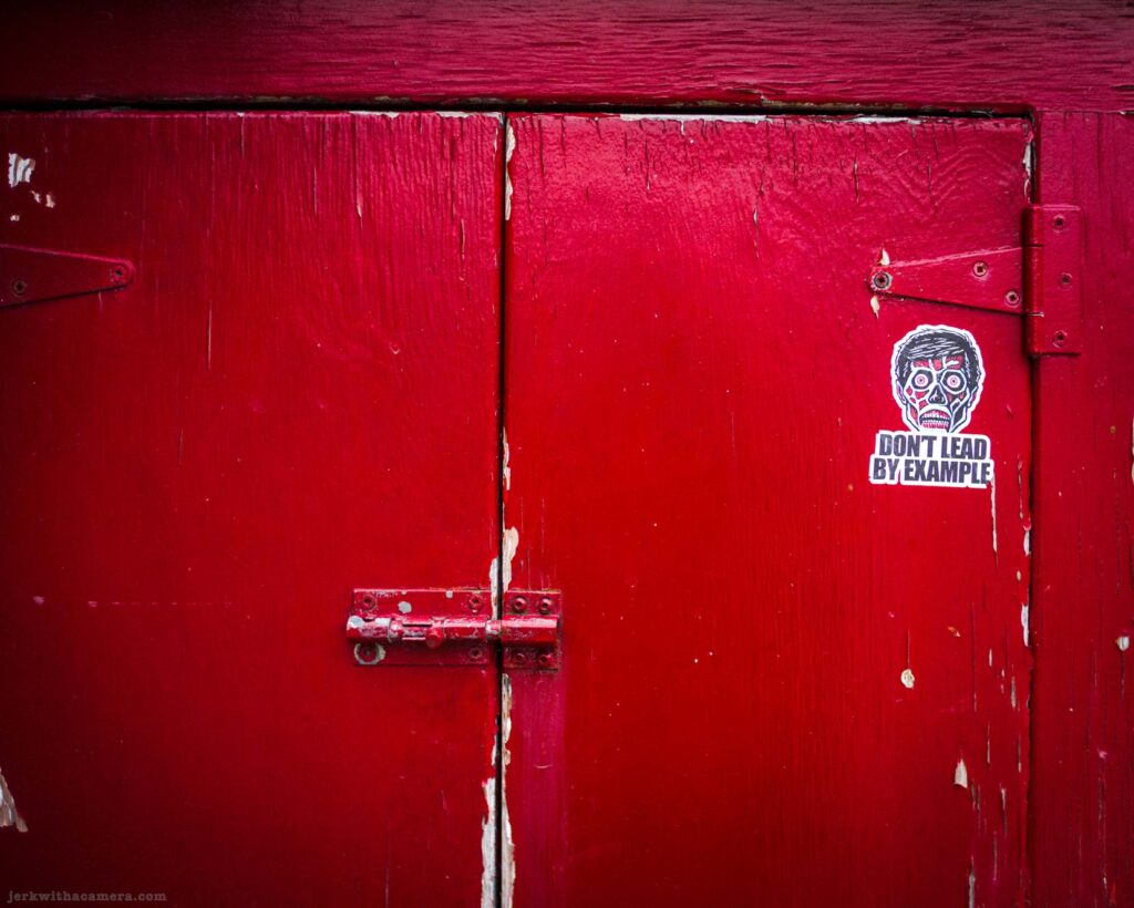 Close-up of red painted wooden double door with heavy peeling paint, metal barrel bolt latch at center, metal hinges, zombie skull sticker reading Don't Lead By Example on right door panel.