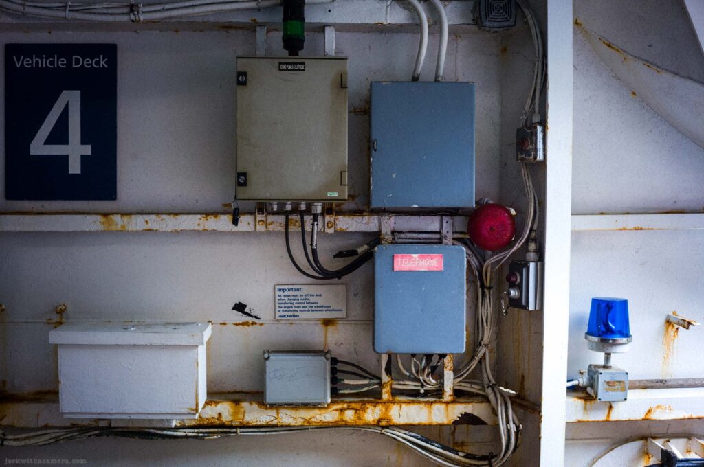 Interior ferry wall with dark blue Vehicle Deck 4 sign, beige Sound Power Telephone box, blue junction box, gray telephone enclosure with red Telephone label, red alarm bell, blue strobe light, conduit runs, rust staining, BCFerries notice.