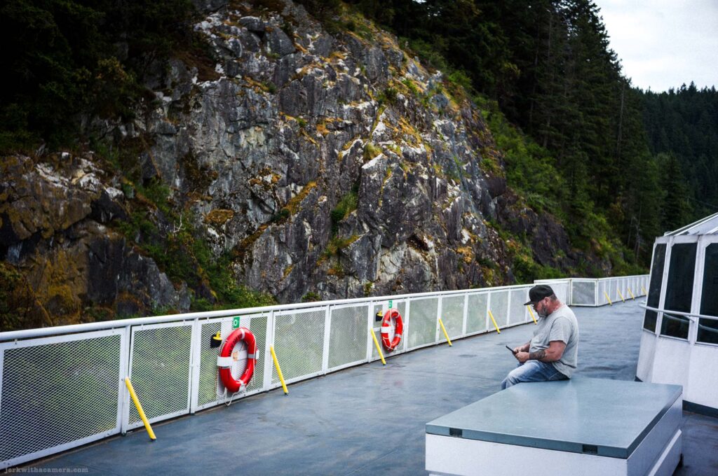Man in gray shirt and jeans seated on gray platform on ferry outer deck, two red and white life rings on white mesh railing, yellow bollards along railing, large moss-covered rocky cliff and conifer forest behind, overcast sky.