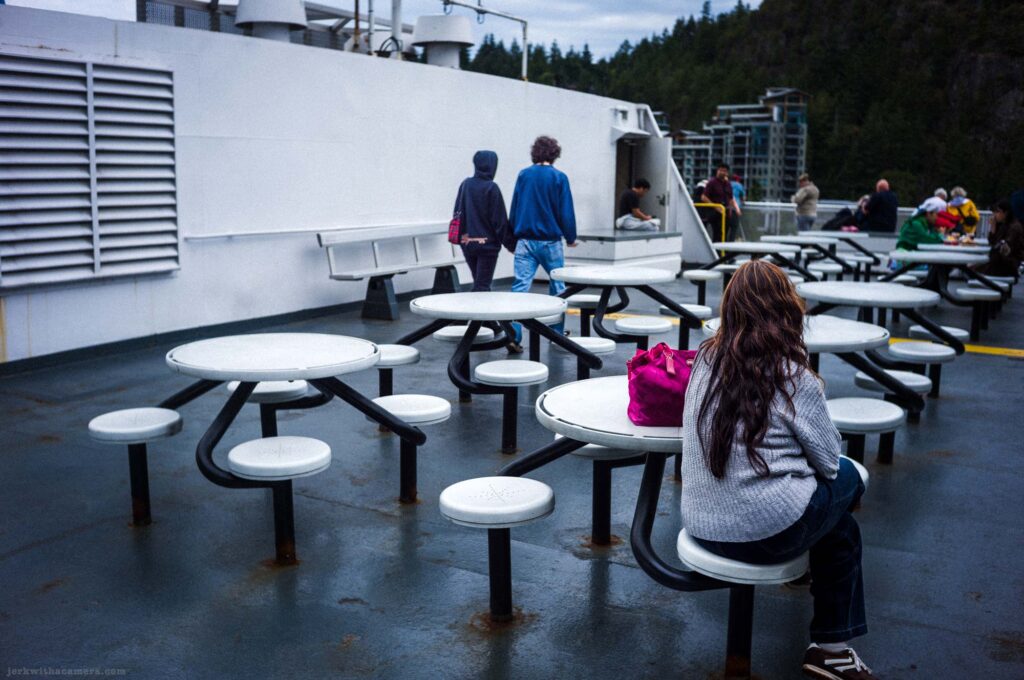Woman with long brown hair in gray sweater at round white table with pink bag in foreground, two people walking toward white superstructure behind, other passengers at tables at right, rocky hillside and scaffolded building in background, overcast.