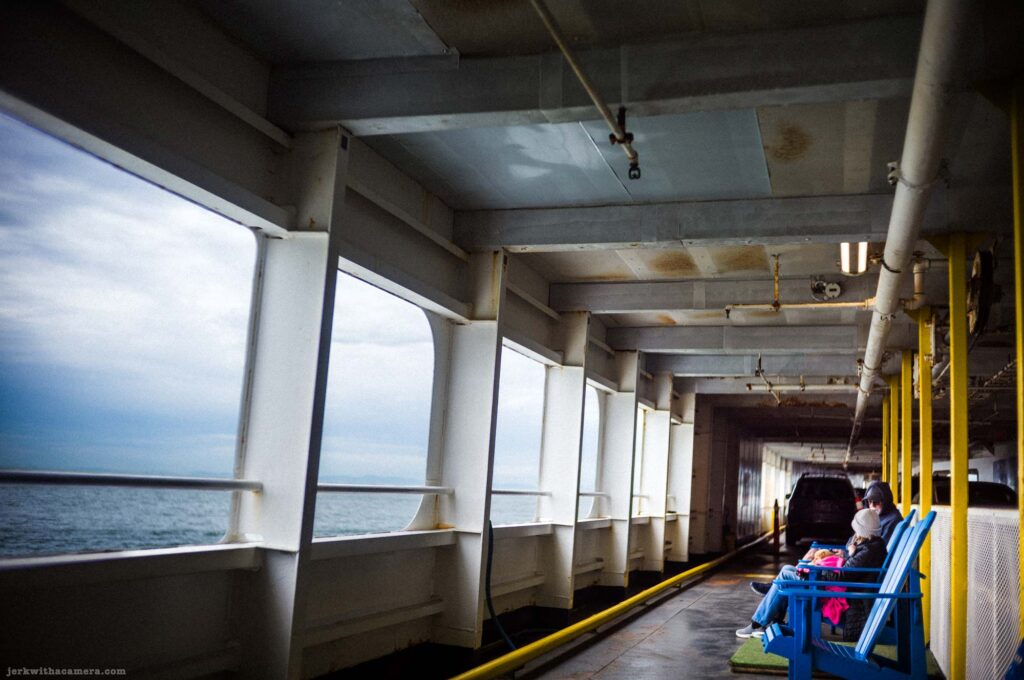 Ferry exterior deck corridor with white structural dividers, yellow support posts, blue Adirondack chairs, a passenger seated at right, and overcast sea visible through openings.