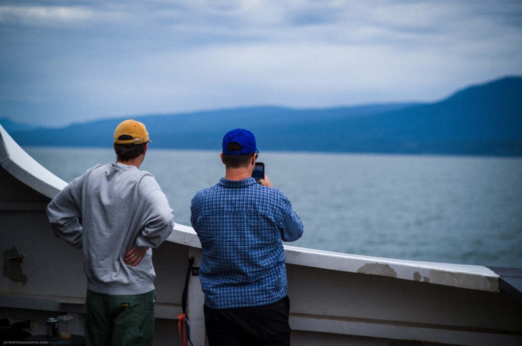 Two men standing at a white painted ferry railing from behind, one in grey hoodie and yellow cap, one in blue plaid shirt and blue cap holding a phone toward the water and mountains.