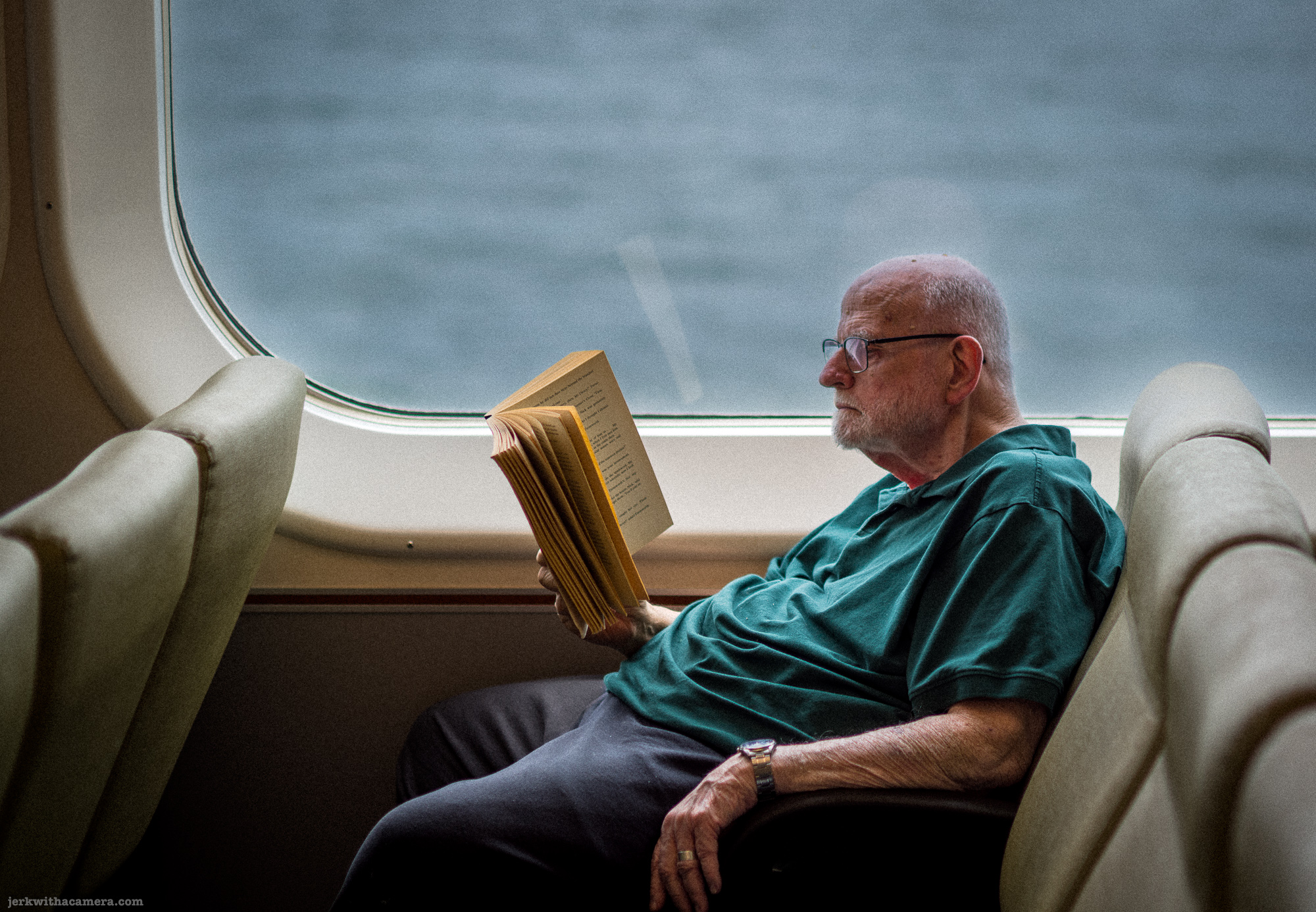 White-haired elderly man with glasses and white beard in a green shirt reading a worn paperback book in a cream upholstered ferry lounge seat, grey water visible through the window.