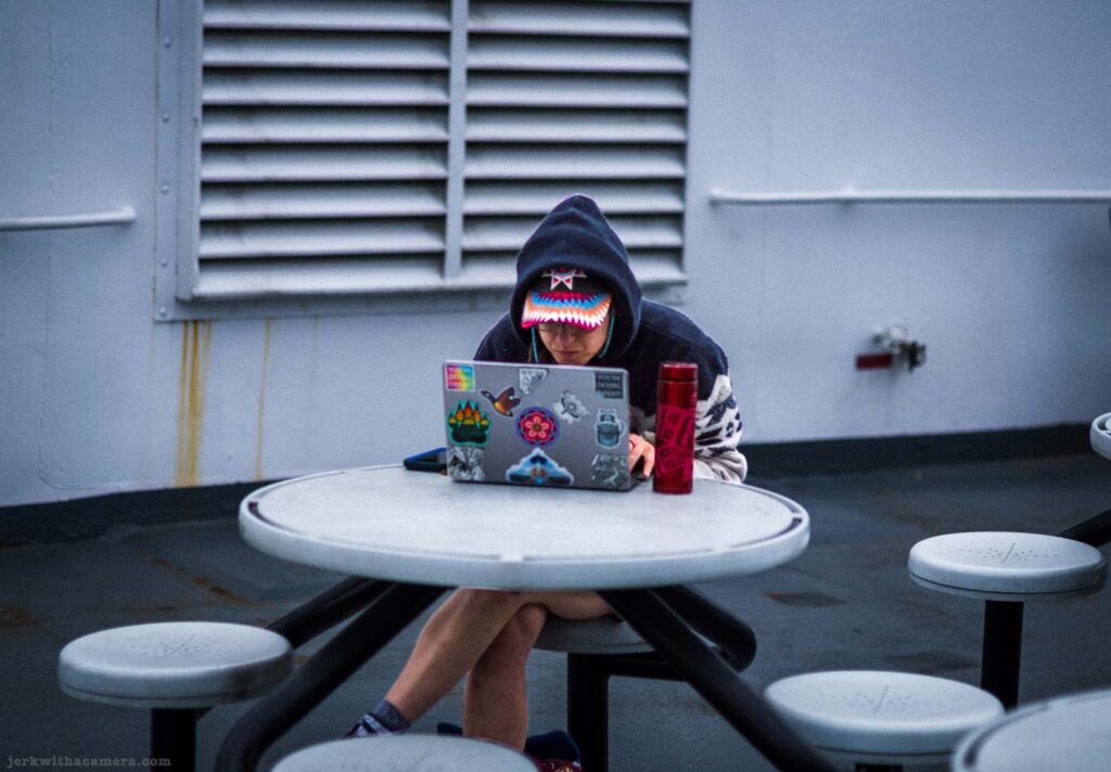 Person in dark hoodie with colourful patterned headband sitting at a round outdoor table on a ferry deck, working on a laptop covered in stickers, red thermos beside them.