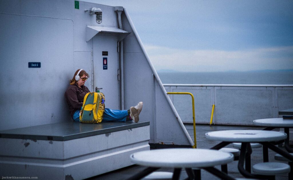 Young woman in brown jacket and blue pants with white headphones and yellow and teal backpack sitting against a white wall on Deck 6 of a ferry, grey sea and horizon visible beyond the railing.