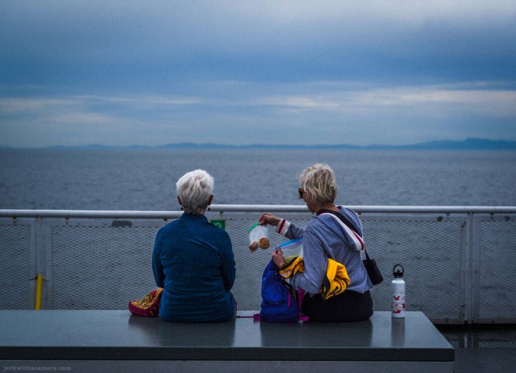 Two older women with short white hair sitting side by side on a grey bench on a ferry deck, one holding an open snack bag, grey sea and distant mountains behind them.