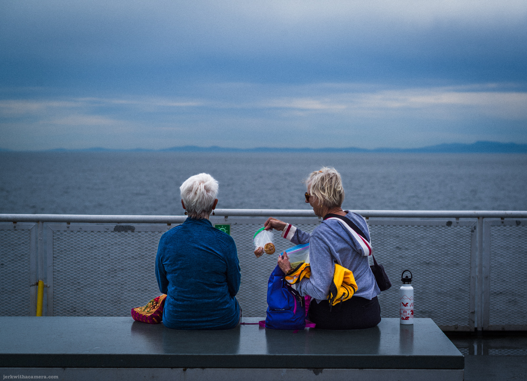 Two older women with short white hair sitting side by side on a grey bench on a ferry deck, one holding an open snack bag, grey sea and distant mountains behind them.
