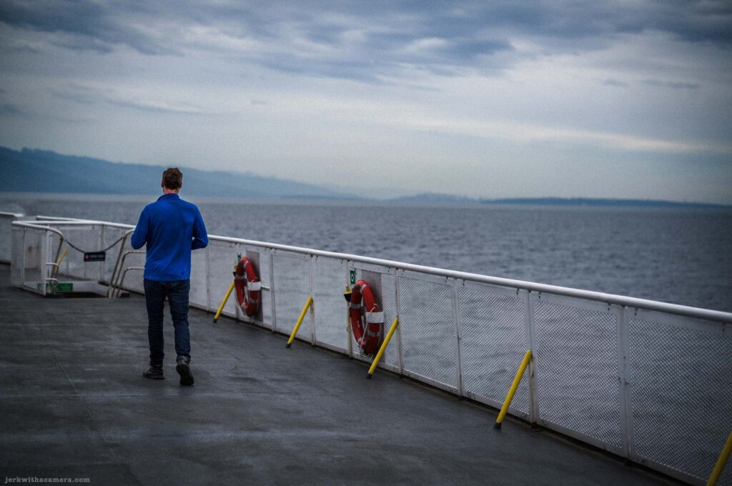 Man in blue jacket and dark jeans walking away from camera on a grey ferry stern deck, white railing with red life rings, grey sea and distant mountains under overcast sky.