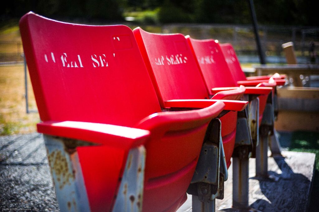 Four red plastic folding stadium seats on rusted white painted brackets from low angle, cut-out lettering including Seal Sec on nearest backrest, gravel ground, chain-link fence and water visible in soft focus background, hard sunlight.