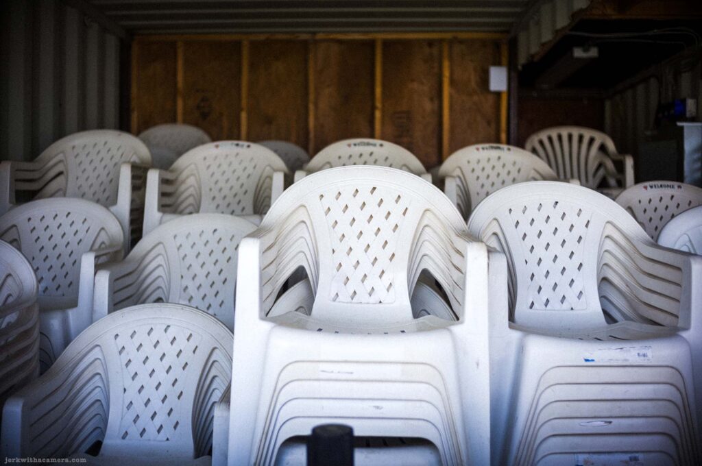 Dense cluster of stacked white plastic armchairs with diamond-pattern backs filling frame, plywood panel background, Welcome logo visible on some chairs, dark interior, ambient light from outside at upper left.