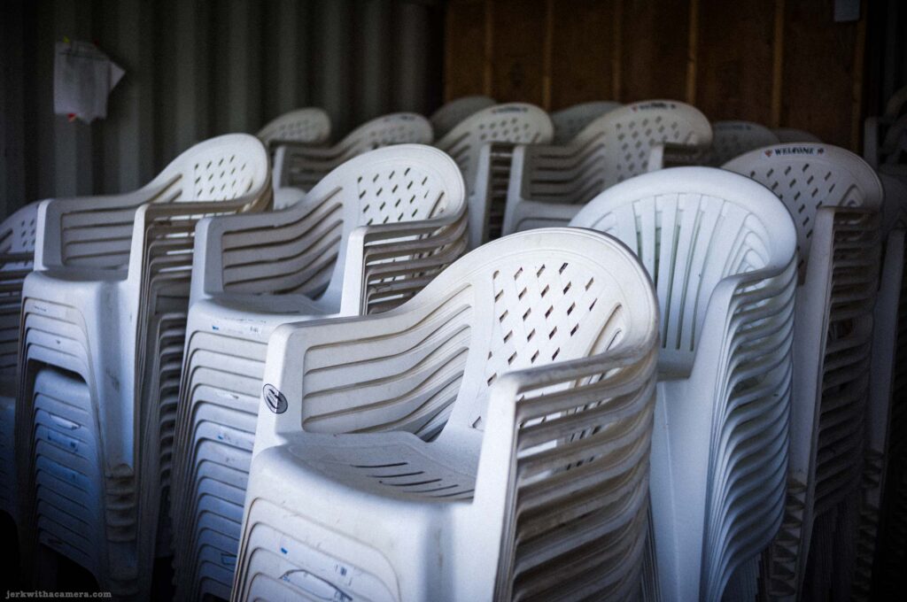 Close view of multiple stacks of white plastic molded armchairs with ribbed and slatted backs in mixed orientation, dark shed interior background, Welcome text visible on at least one chair.