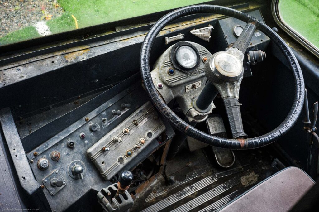 Looking through windshield into old vehicle interior, large black steering wheel with wrapped rim, deteriorated cast metal dashboard hub with small gauge, toggle switches and colored indicator lights, cast metal switch panel, gear lever, passenger seat at right, green lawn outside.