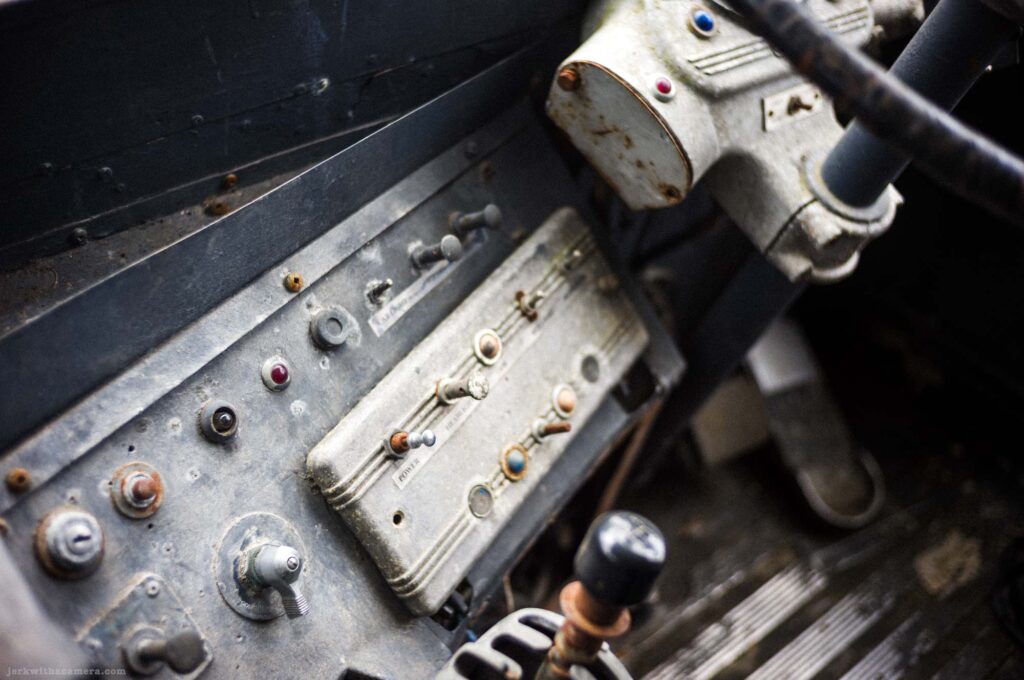 Close-up of worn cast metal vehicle dashboard panel with multiple toggle switches, colored indicator lights in red blue and orange, rubber conduit, portion of steering wheel at upper right, Power label visible on switch panel.