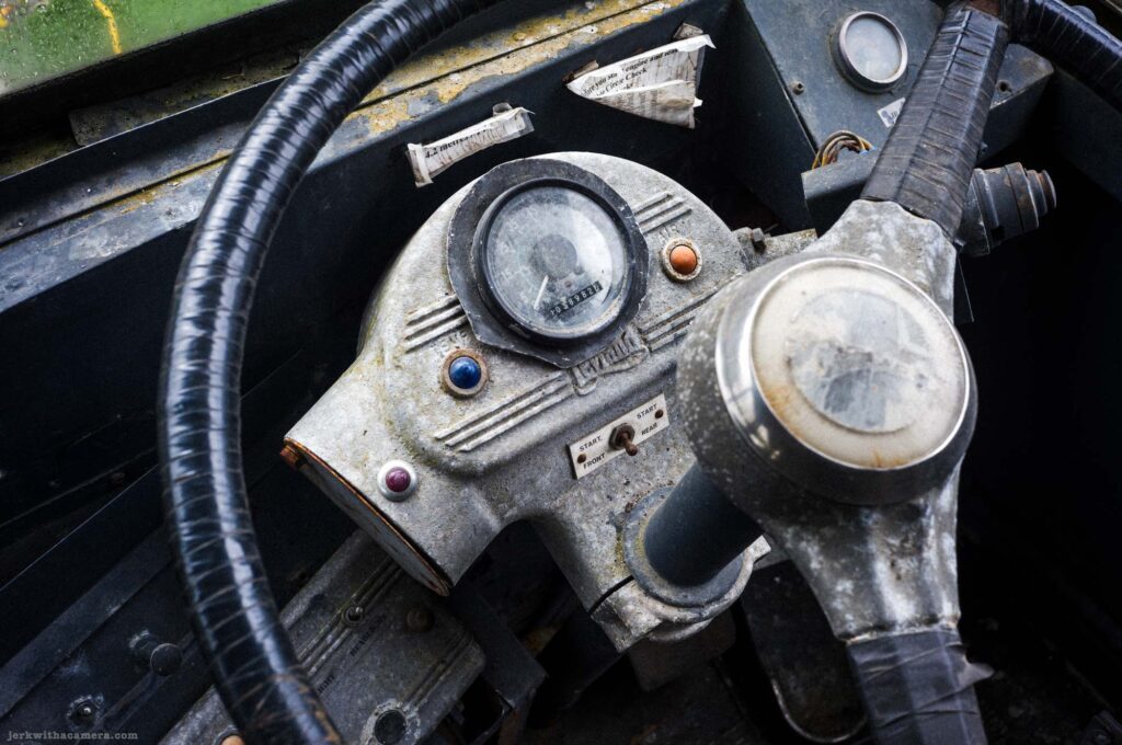 Close-up of vintage steering wheel center hub, round odometer in black housing, small colored indicator lights in blue orange and red, cast metal housing with decorative ribbing, Start Front Rear labels, rubber-wrapped steering column visible.