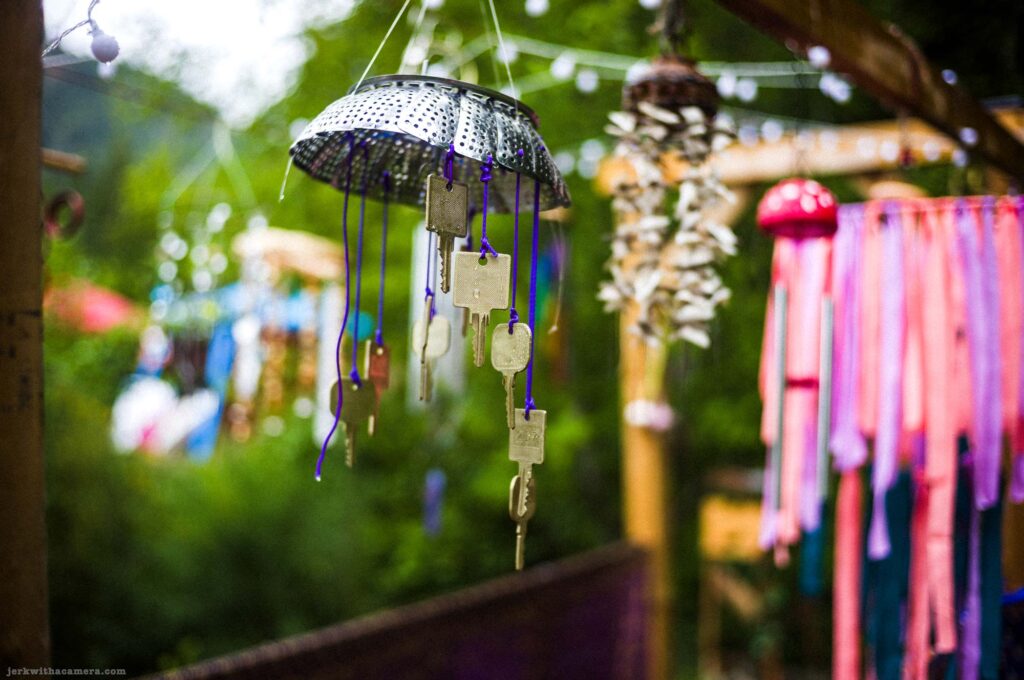 Wind chime made from inverted stainless steel colander with multiple brass and silver keys on purple cord, outdoor setting, blurred colorful hanging decorations in background including shell mobile and ribbon chime, green foliage behind.