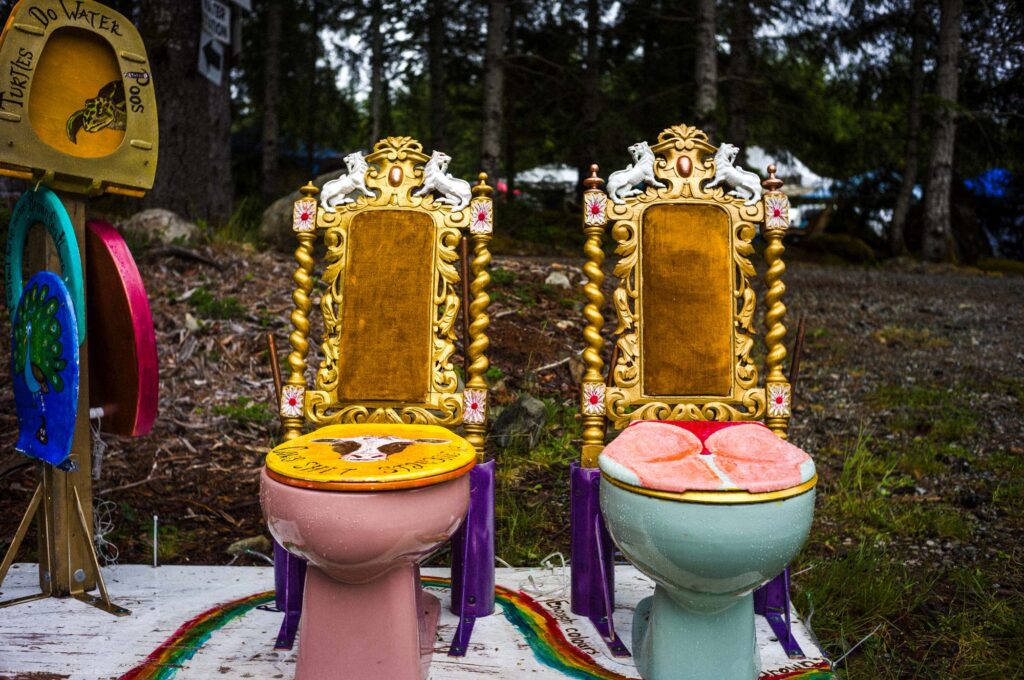 Pink toilet with painted cow lid and gold baroque chair back topped with white horse figures at left, blue toilet with lip-shaped pink lid and matching gold back at right, rainbow painted base, painted toilet lids on stands at left, trees and overcast sky behind.