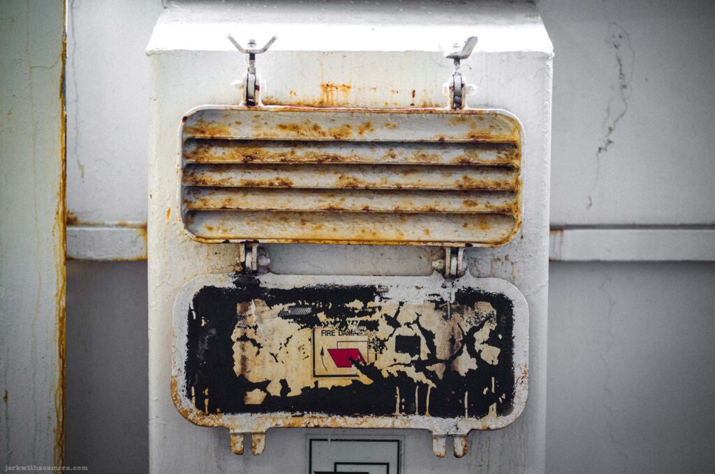 White painted metal fire damper housing face-on, rusted hinged louvered grille on top, deteriorated black and white label reading Fire Damp with small red graphic below, wing-nut fasteners at top corners, white wall and structural column around unit.