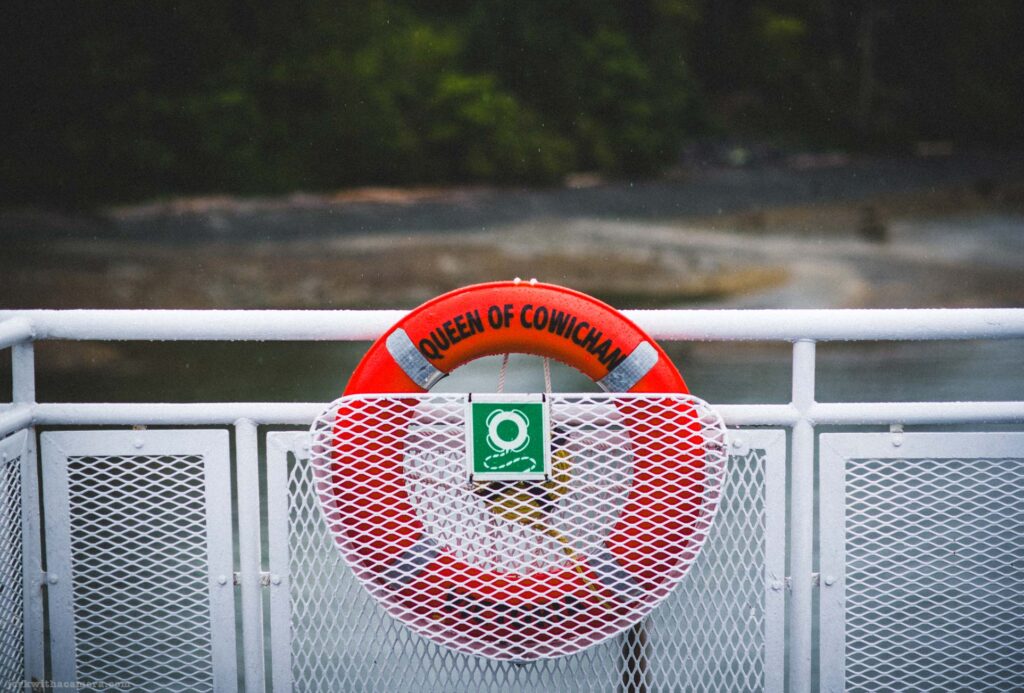 Orange life ring with black text reading Queen of Cowichan and two reflective silver bands, mounted in white mesh railing bracket with green safety sign showing life ring icon, white metal railing, rocky shoreline and water behind, rain visible.