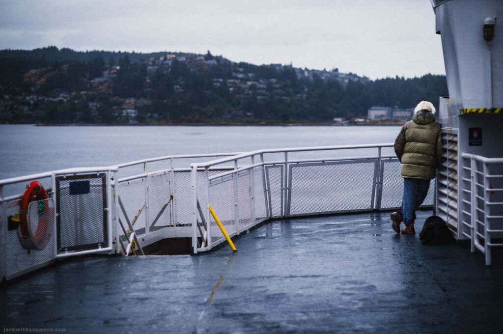 Person with white-blond hair in olive green hooded puffer jacket and jeans at end of ferry outer deck, leaning against white superstructure, black bag at feet, life ring on railing at left, hillside with houses across gray water, overcast sky.