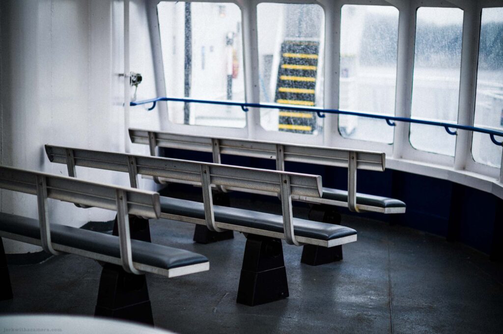 Interior ferry curved window alcove, two empty metal-framed benches with black vinyl seats facing large windows, blue tubular handrail along window, staircase with yellow anti-slip treads visible through rain-spattered glass outside, dark floor.