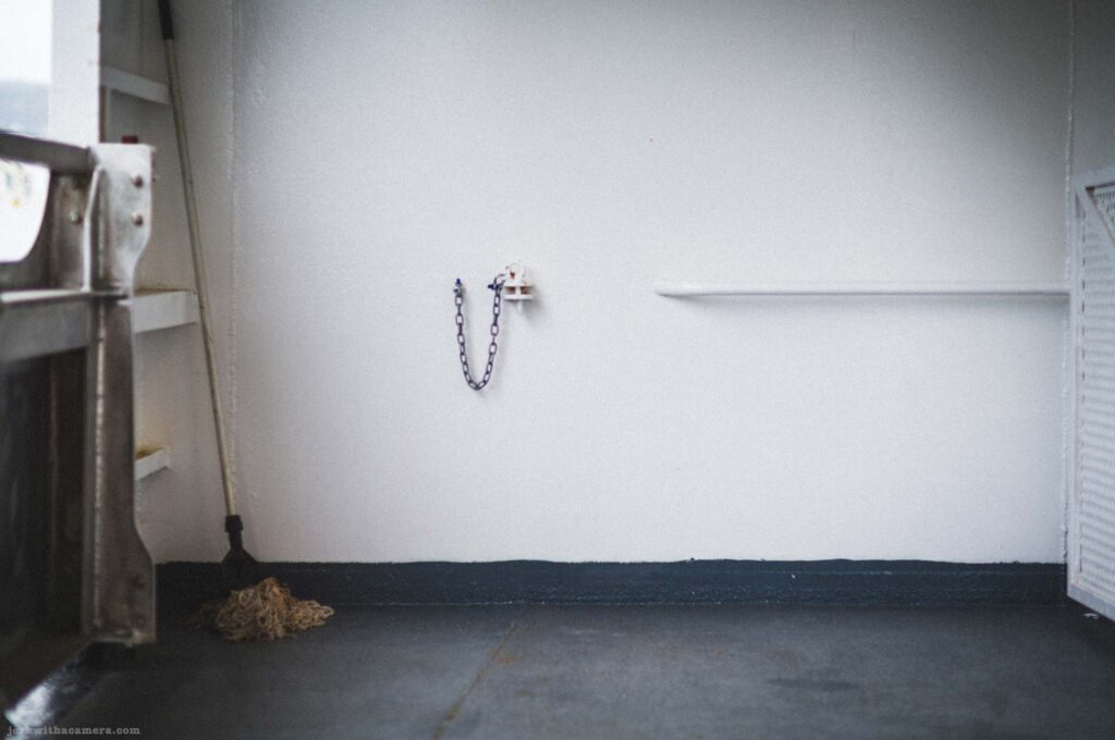 White painted interior ferry wall, short metal chain looped in U-shape between two mounted anchor points, horizontal white ledge at right, mop with natural fiber head leaning at lower left against metal bracket, dark floor at base.