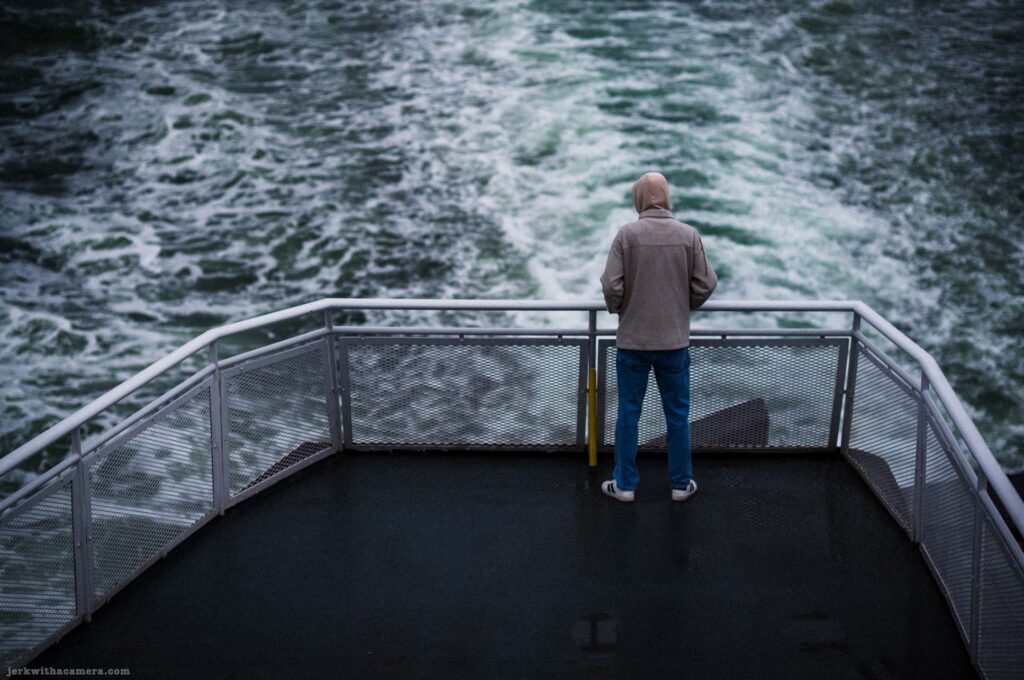 Person in gray zip-up and blue jeans standing centered at ferry stern, symmetric V-shaped white railing framing figure from above, large churning white and green wake filling background, wet dark deck in lower half.
