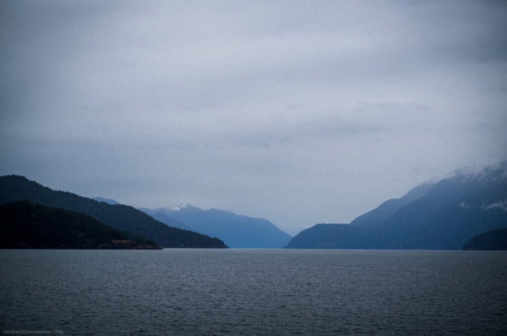 Dark blue-gray calm water in lower half, layered mountain ridges flanking a fjord channel receding into distance, snow-capped peaks on right range, low cloud and mist at mountain tops, heavy overcast sky.