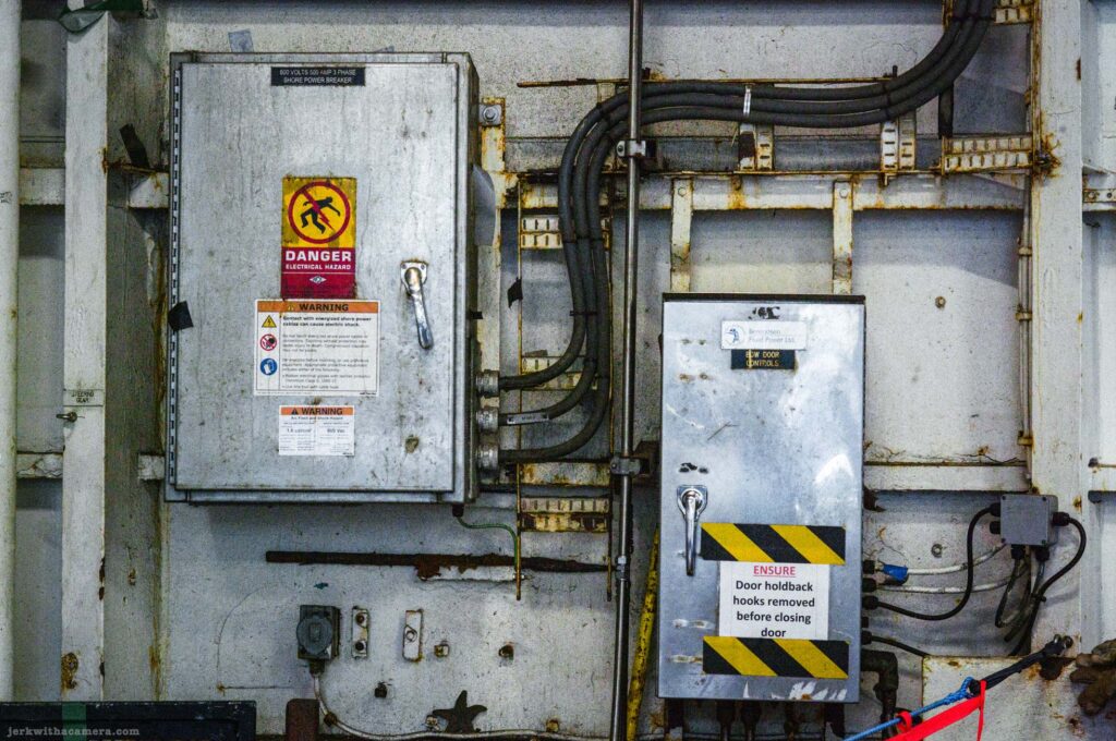 Two metal enclosures on rusted white wall, left labeled 800 Volts 500 Amp 3 Phase Shore Power Breaker with Danger Electrical Hazard and Warning signs, right labeled Bow Door Controls by Bennson Fluid Power with hazard tape and ensure door holdback hooks sign, conduit runs, rust throughout.