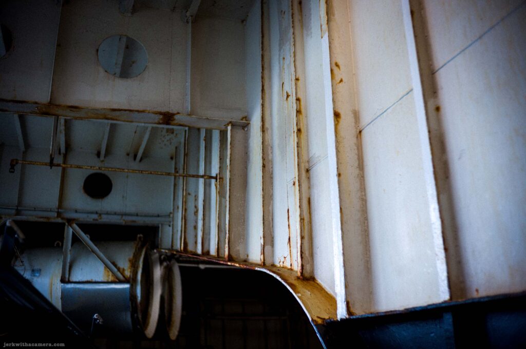 Upward oblique view of rusted white painted steel panels and structural ribs in ferry vehicle deck, two circular porthole openings in left panel, metal railing grid, curved vehicle door or ramp structure at lower right.