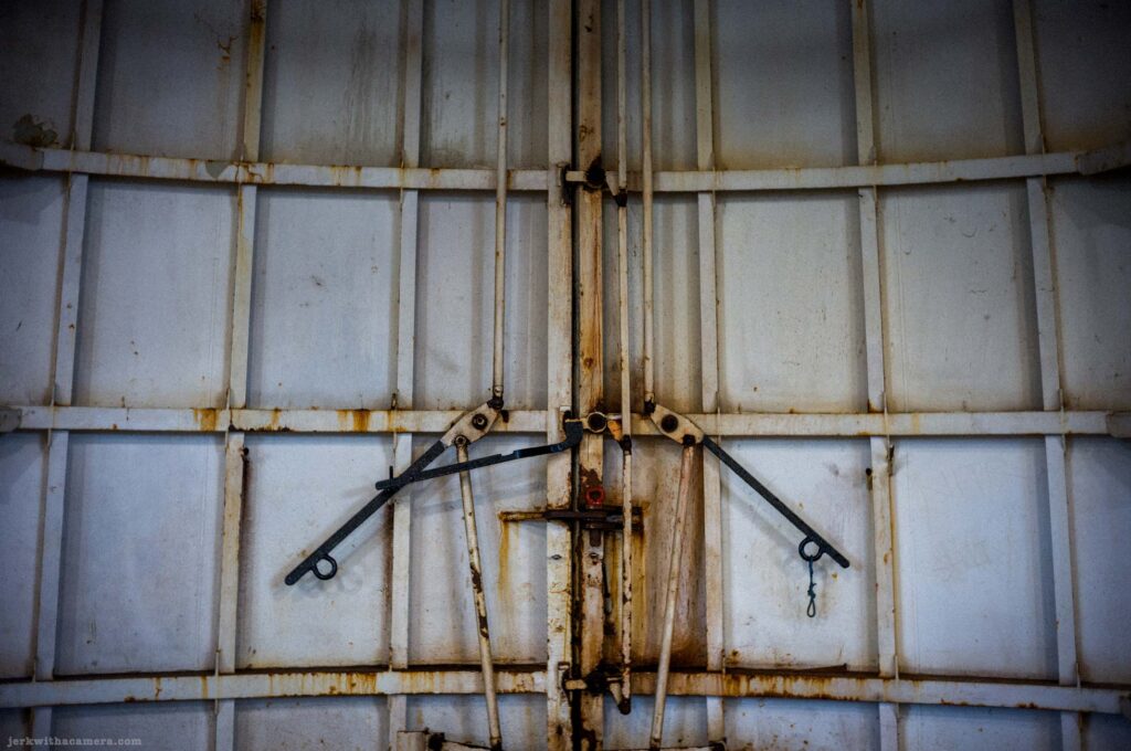 White painted corrugated ferry bow door panels, central locking mechanism with two black metal lever arms extending symmetrically from pivot hardware, rust staining along seams and joints, red fitting at pivot, small wire loop at lower right.