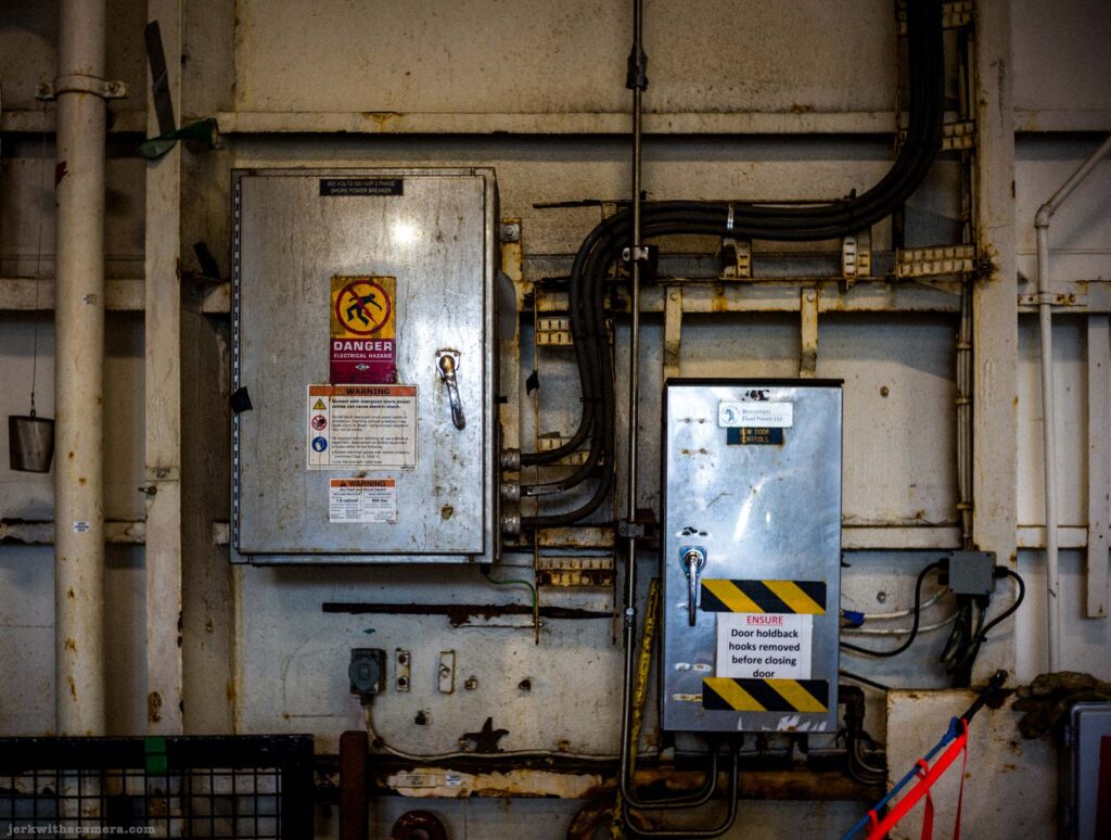 Wider view of rusted BC Ferry vehicle deck wall, left enclosure with Danger Electrical Hazard and shore power breaker label and Warning signs, right with Bow Door Controls label with hazard tape and door holdback hooks sign, conduit runs, pipes above, rust throughout.