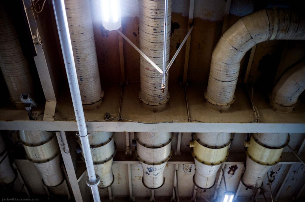 Looking up at cluster of large cream-wrapped cylindrical duct pipes running vertically and turning horizontally, metal grid framework separating levels, fluorescent tube light between center pipes, some pipes with yellow-green collar fittings at lower level.