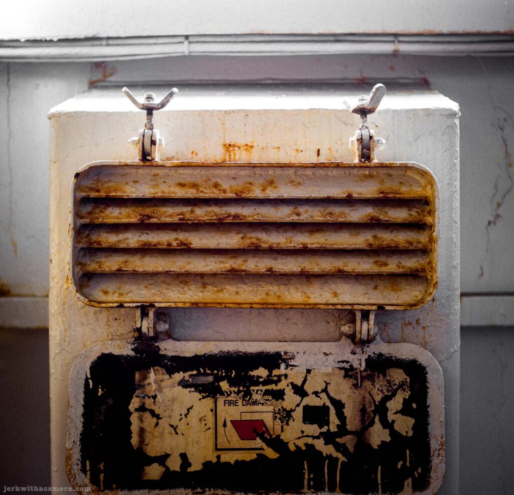 Square white-painted metal fire damper unit face-on, hinged louvered grille heavily rusted, Fire Damp label with small red graphic mostly peeled on black backing below, wing-nut fasteners at top corners, white wall and dark floor around unit.