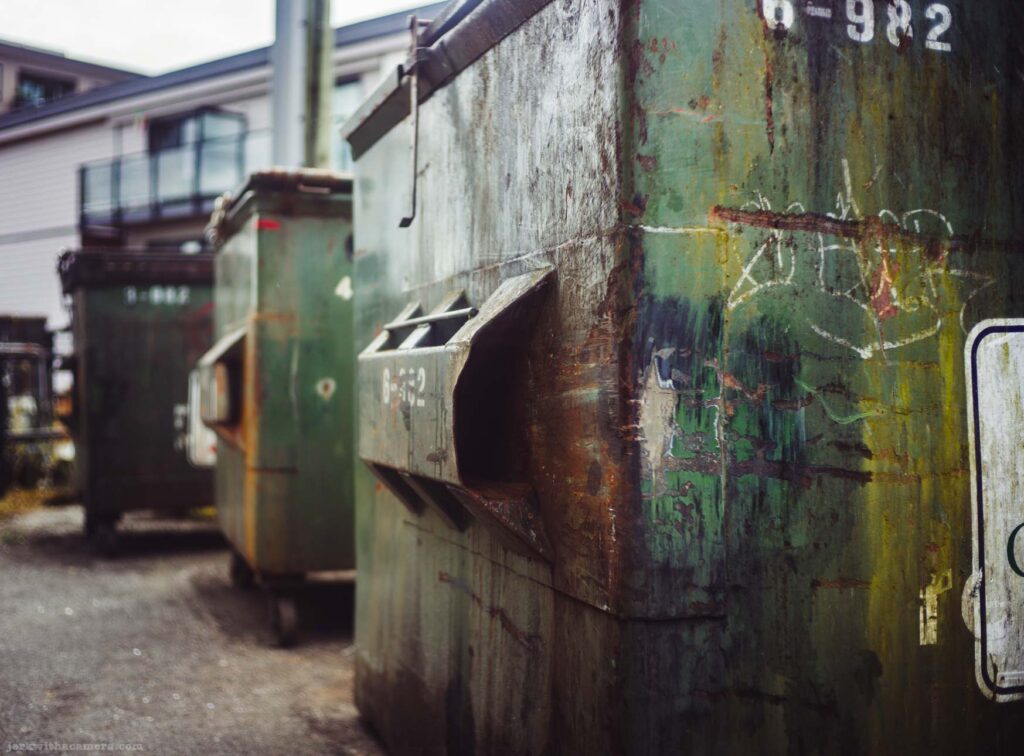 Wide view of a row of green metal dumpsters stenciled 6-982 in a parking area behind an apartment building.