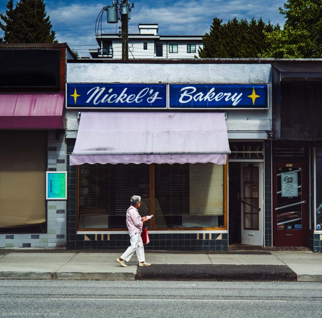 A person in white walking right to left past Nickel's Bakery, red sports car partially visible at right.