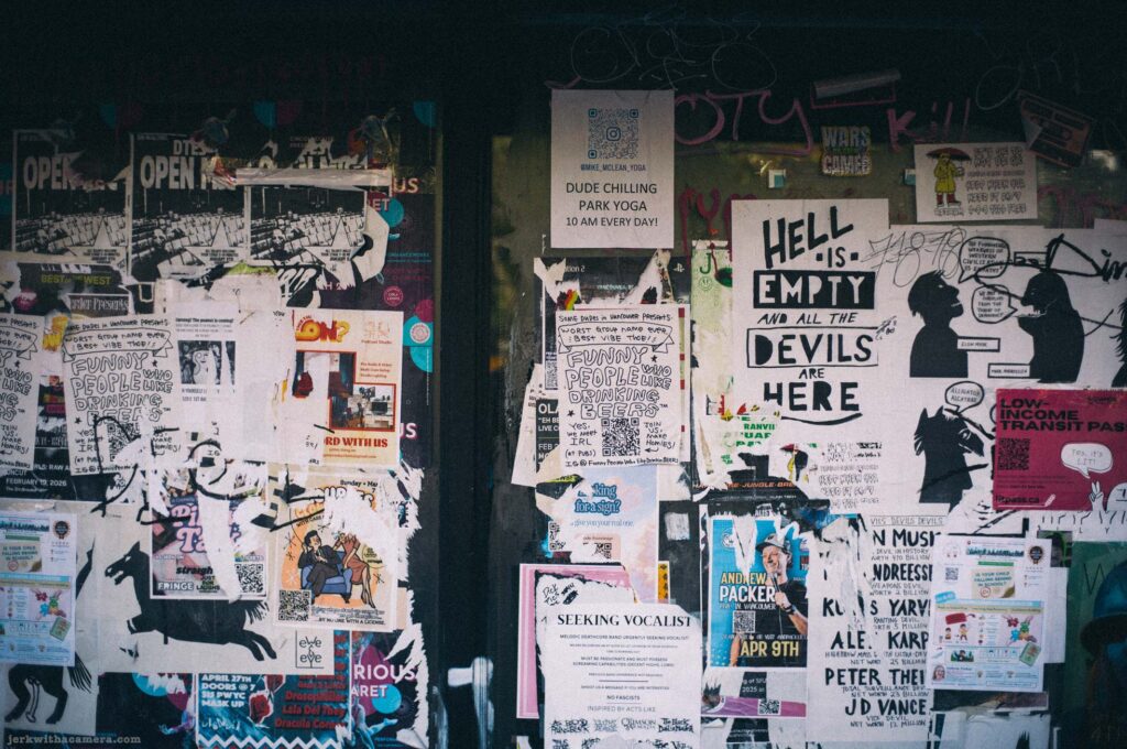 Wall densely covered in overlapping flyers and posters. Central screen-printed poster reads: Hell Is Empty and All the Devils Are Here.