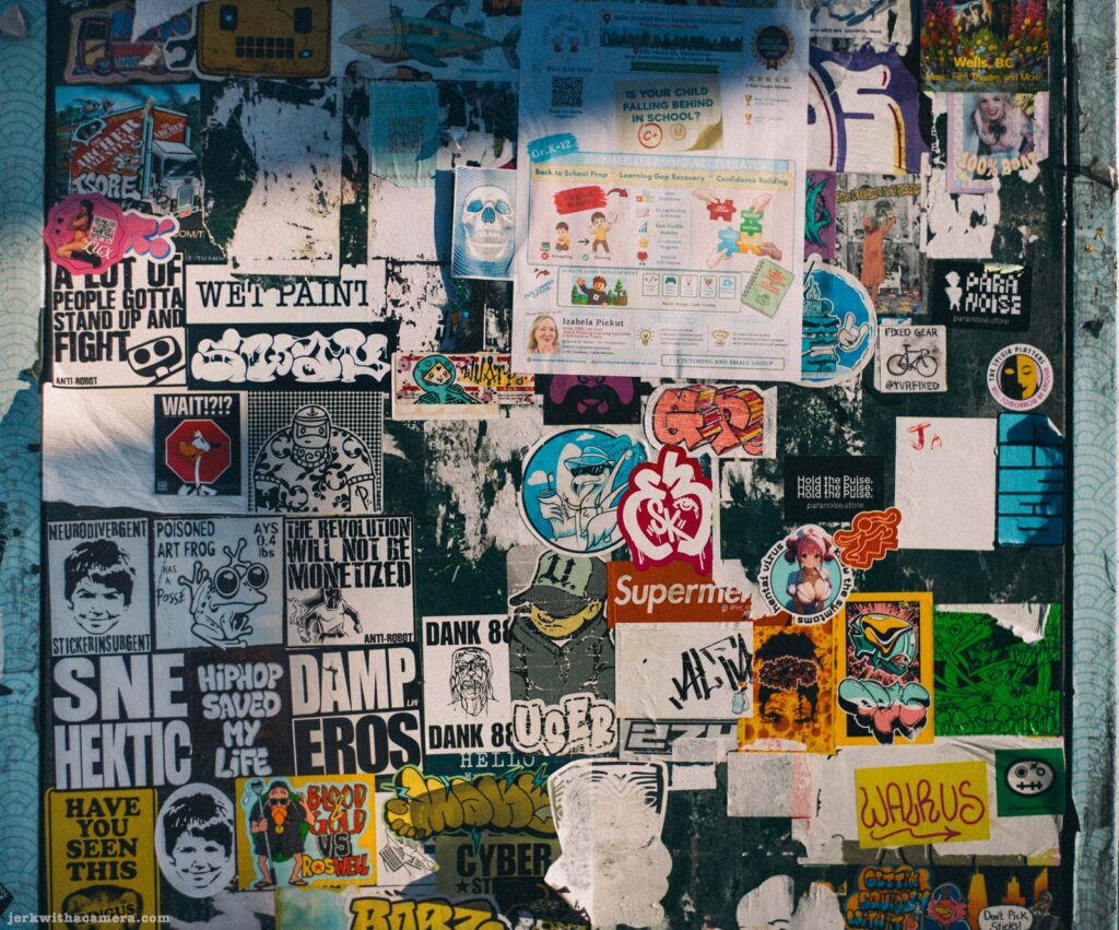 Grey utility box completely covered in layered stickers and paste-up flyers. Readable text includes Wet Paint, SNE Hektic, Damp Heros, Supermen.