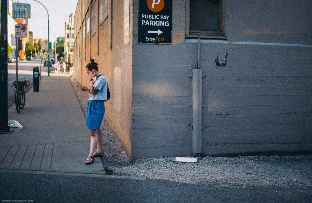 A woman stands on a sidewalk looking at her phone beside a public pay parking sign. EasyPark branding on the sign.