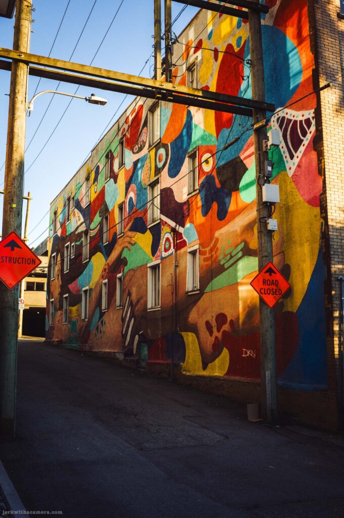 Large multi-coloured abstract mural covers the full side of a building. Two orange construction signs at the base of the alley.