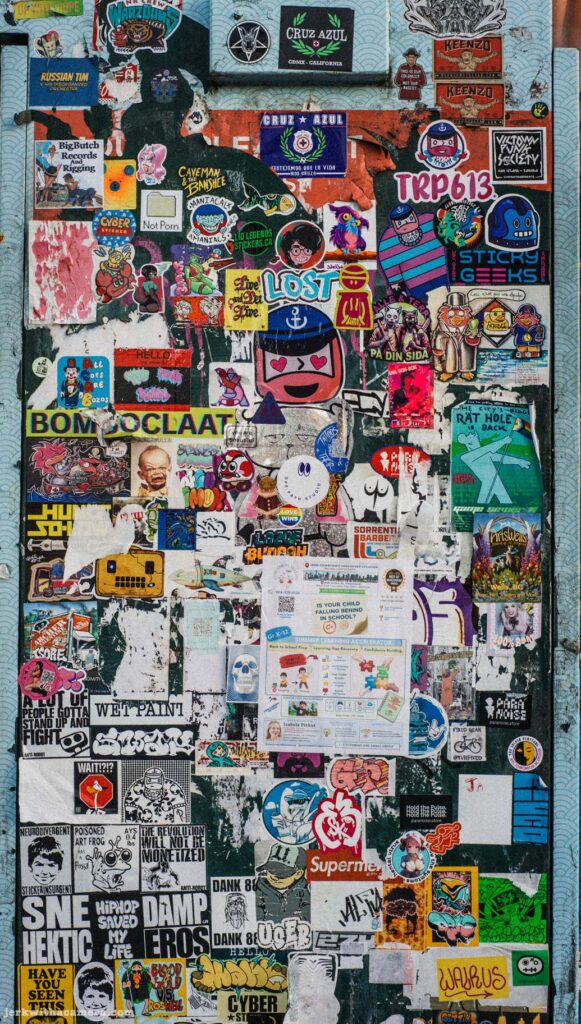 Tall utility box covered in stickers. Bom Oclaaf, TRP613, Lost visible. Person partially visible at left edge.