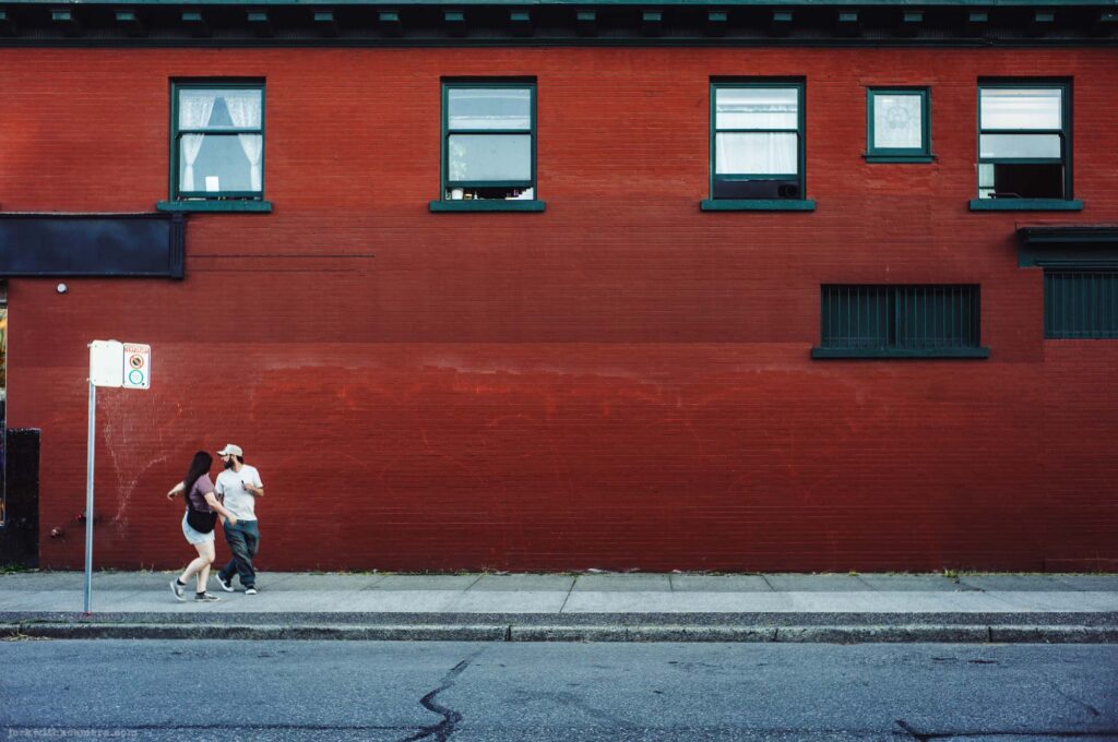Two people walk along a sidewalk past a large red painted brick building. Street sign pole visible at left.