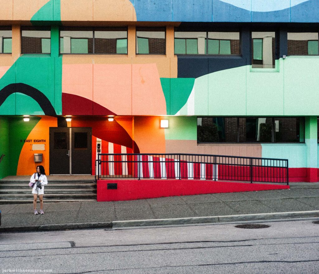 Multi-coloured school building facade with a large abstract mural. A person with a backpack stands at the entrance ramp.