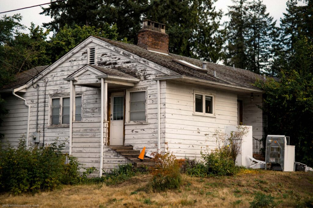 A small white house with peeling paint, a front porch, overgrown vegetation, and utility equipment beside the building.