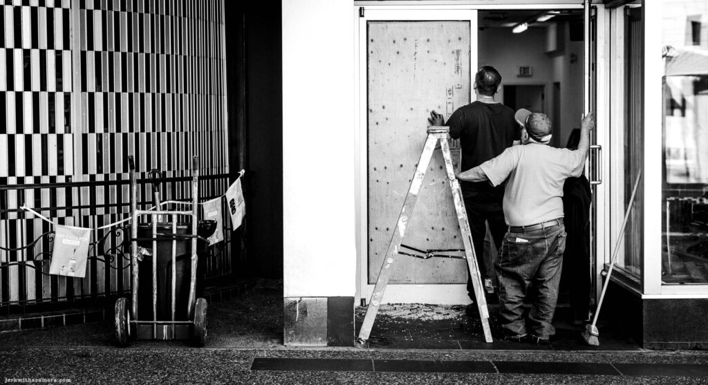 Two workers install a plywood board over a storefront doorway. A ladder leans against the frame and a hand trolley with bags sits to the left.