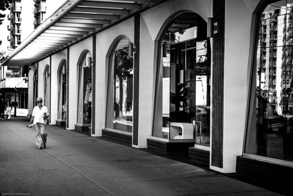 A woman in a white blouse walks along a sidewalk past a store with arched windows. A CB2 sign is above the entrance.