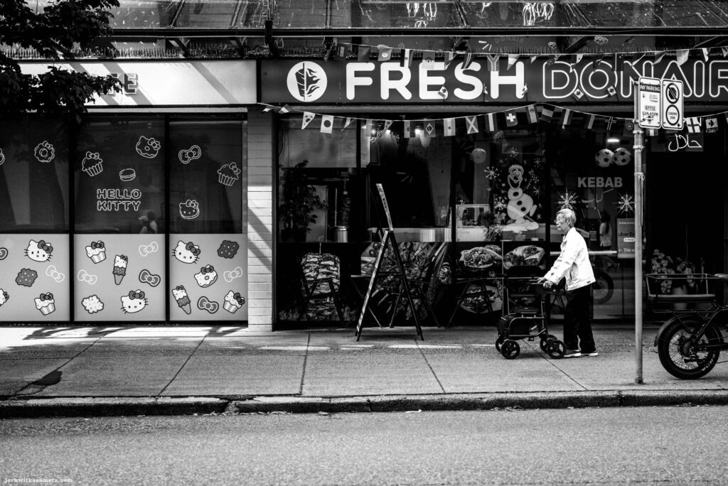 An elderly person in a white top pushes a rollator walker along the sidewalk in front of the Fresh Donair restaurant. The window next door is covered with large Hello Kitty character graphics.