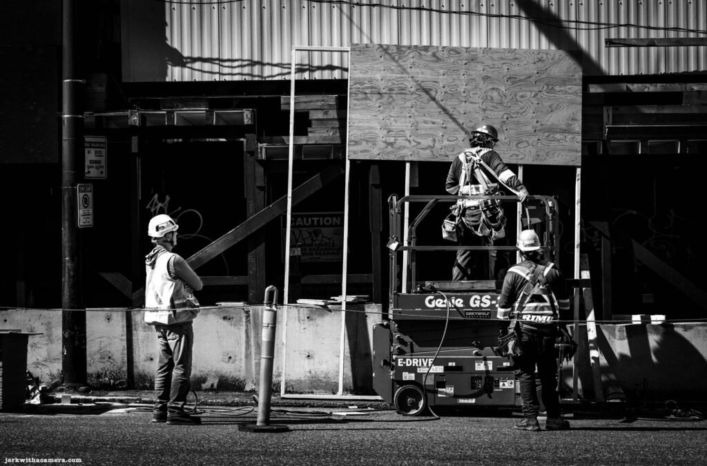 Three construction workers in high-visibility vests and hard hats at a building site. One stands watching, one is on an elevated scissor lift, and a third stands at the base.